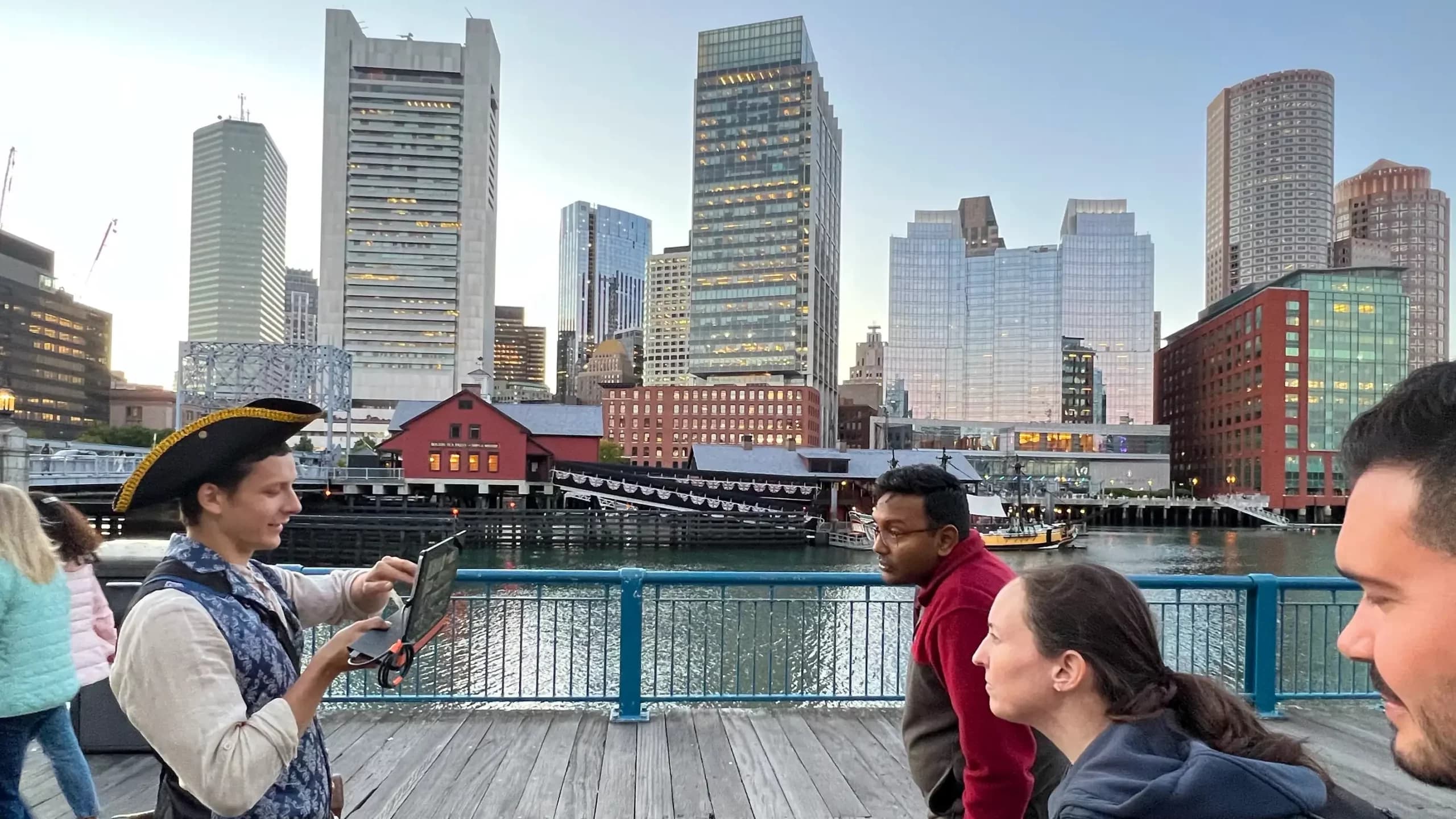 A guide in a tricorn hat holds up a small screen on the Congress Street bridge at dusk, showing a group archival views with the Boston Tea Party Ships and the downtown skyline behind