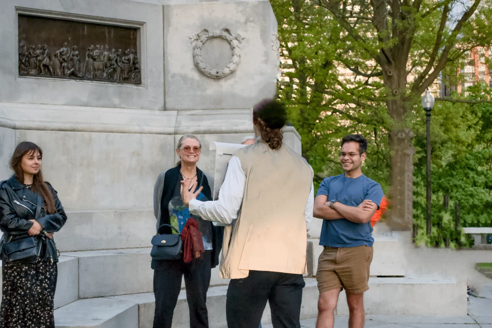 A Beaming guide holding a rolled map, mid-story with three smiling guests beside a bronze relief panel on a Boston monument